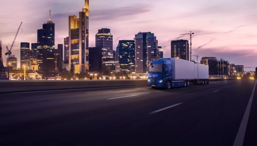 Elektryczna ciężarówka Renault po zachodzie słońca jedzie drogą, w tle widać duże biurowce miasta