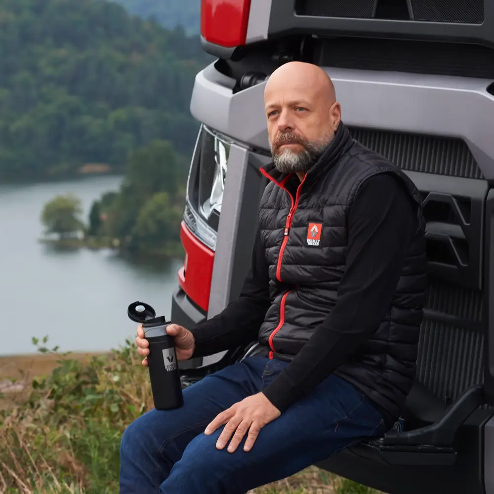 mężczyzna ubrany w kamizelkę Renault Trucks siedzący na zderzaku swojej ciężarówki gdzieś w plenerze. W ręku trzyma termos z logiem Renault Trucks