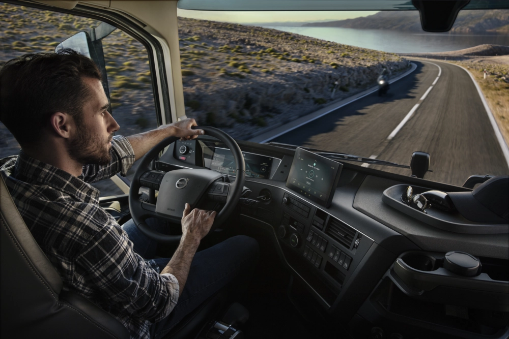 Kierowca ciężarówki Volvo siedzący w kabinie przez przednią szybę widać drogę oraz fjord w tle.